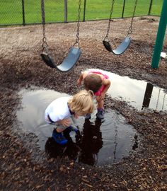 two children playing in a puddle at a playground with swings and water on the ground