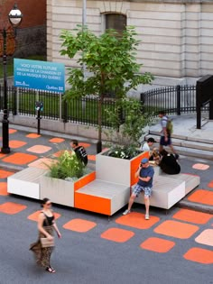 people are walking on the sidewalk near an orange and white bench with plants in it