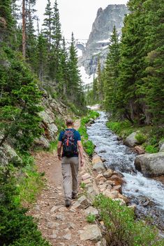 a man with a backpack walks along a rocky path next to a stream and trees