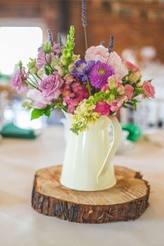 a white pitcher filled with flowers sitting on top of a wooden slice at a table