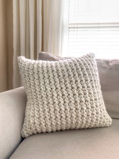 a white knitted pillow sitting on top of a gray couch next to a window