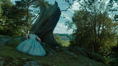 a woman in a blue dress sitting next to a large tree on top of a hill