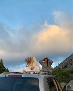 two people sitting on the roof of a car in front of a mountain with clouds