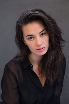 a woman with long dark hair and black shirt posing for a photo in front of a gray background