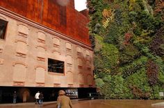 people are walking around in front of a building with green walls and plants growing on it