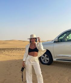 a woman standing in the sand with a cell phone to her ear and a car behind her