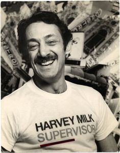 a smiling man wearing a harvey milk supervisor t - shirt in front of a pile of junk