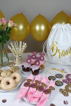 a table topped with donuts and cupcakes covered in pink frosting next to gold balloons