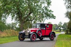 an old red car is driving down the road in front of some trees and grass