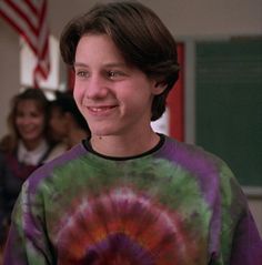 a young boy wearing a tie - dyed shirt smiles at the camera while standing in front of an american flag