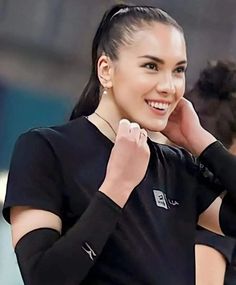 a woman in black shirt smiling and holding her hand up to her ear with both hands