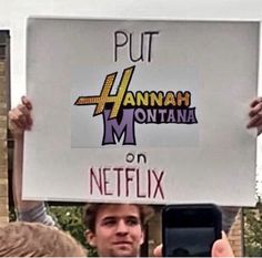 a man holding up a sign that reads put hannah montana on netflixx and holds his cell phone in front of him