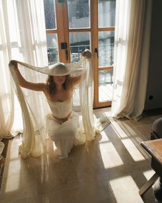 a woman sitting on the floor in front of a window wearing a white dress and hat