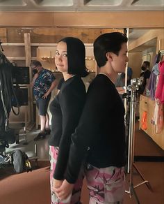 two women standing next to each other in front of a mirror with clothes hanging on the wall