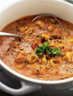 a white bowl filled with pasta and meat soup on top of a wooden table next to a spoon