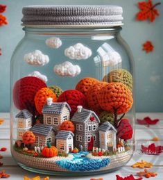 a glass jar filled with crochet houses and trees in the fall, surrounded by falling leaves