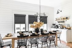 the dining room table is set with black chairs and white walls, along with two windows that have roman shades on them