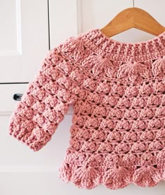 a pink crocheted sweater hanging on a wooden hanger in front of white cupboards