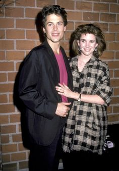 a man and woman standing next to each other in front of a brick wall with their arms around each other