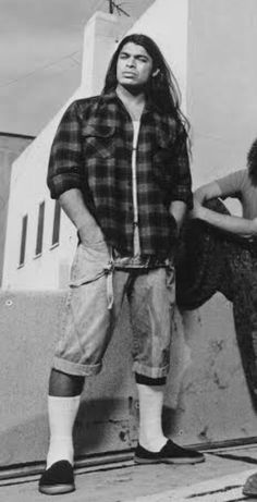 a man with long hair standing next to a wall and holding a skateboard in his hands