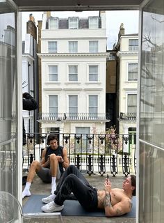 two men are sitting on the ground in front of a building and one man is laying down