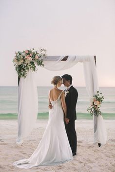 a bride and groom standing on the beach under an arch decorated with floral arrangements for their wedding ceremony