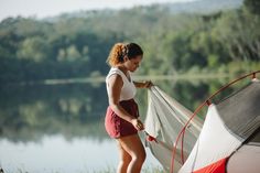 a woman standing next to a red and white tent near a body of water with trees in the background