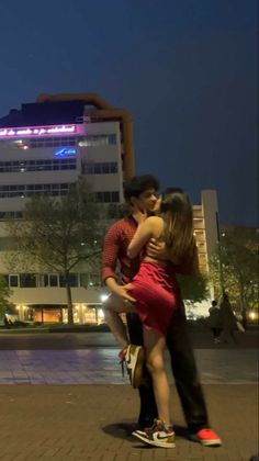 a man and woman hugging in front of a building at night with the lights on