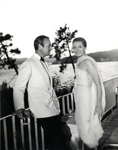 a man and woman standing next to each other in front of the ocean with trees