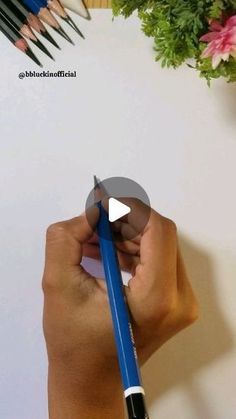 a person holding a blue pencil in their right hand and writing on the paper with crayons next to them