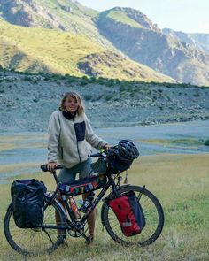 a woman is standing next to her bike in the grass with mountains in the background