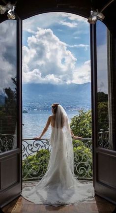 a woman in a wedding dress is looking out an open door at the water and mountains