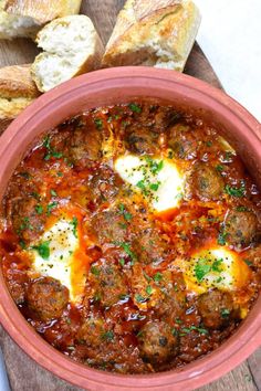 a bowl filled with meatballs and cheese on top of a wooden table next to bread