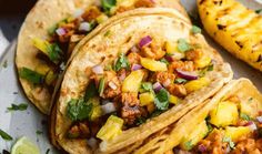 three tacos on a plate with pineapples and cilantro in the background