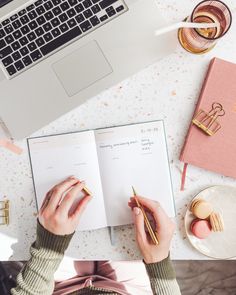 a woman is writing on her notebook while sitting at a desk with a laptop and other office supplies