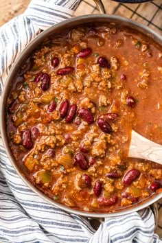 a large pot filled with chili and beans on top of a wooden table next to a blue towel