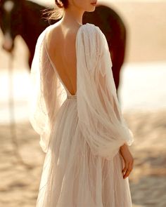 a woman in a white dress standing next to a brown horse on the beach with her back turned