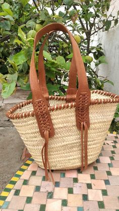 a woven basket with leather handles and tassels sitting on a tiled floor in front of some bushes