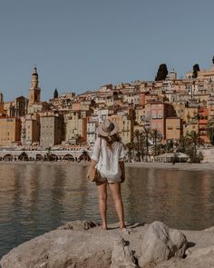 a woman standing on top of a rock next to the ocean with buildings in the background