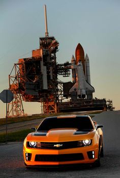 an orange sports car parked in front of a rocket ship