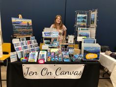 a woman standing behind a table with paintings and cards on it that says ten on canvass