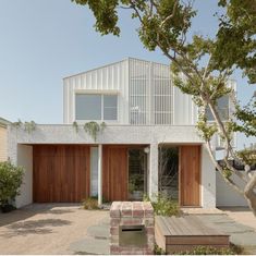 a white house with wooden garage doors and brick steps leading to the front door is surrounded by greenery