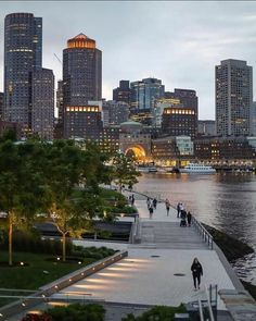 people are walking along the water in front of some tall buildings at dusk, with lights on