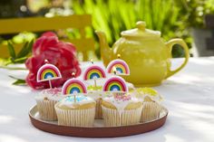 cupcakes with rainbow decorations on a plate next to a teapot and flowers