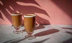 two wine glasses sitting on top of a table next to a pink wall and shadow