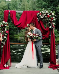 a bride and groom kissing in front of an outdoor ceremony arch with red drapes