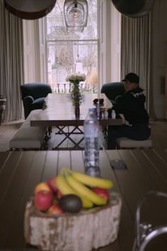 a wooden table topped with fruit next to a vase filled with bananas and apples on top of a hard wood floor