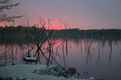 two people sitting on a bench in front of a lake at sunset with trees reflected in the water