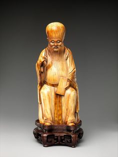 a carved wooden figure of an old man sitting on a stand with his hands in his pockets