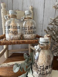 three glass bottles with christmas message on them sitting next to a wooden chair and tree